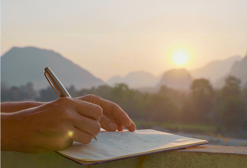 Hände schreiben in ein Journal vor Sonnenuntergang über Bergketten